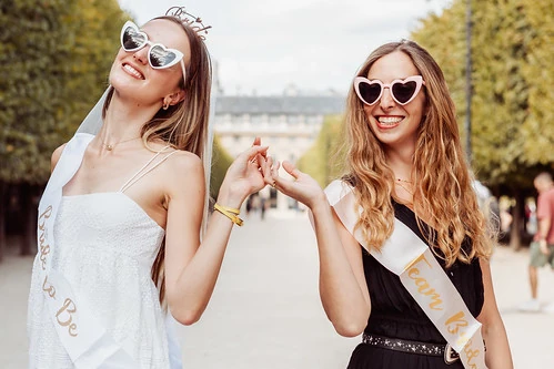 Deux amies portant des lunettes en cœur et des écharpes EVJF posent joyeusement dans un parc 