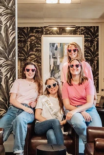 Quatre amies souriantes portant des lunettes en forme de cœur posent ensemble dans un salon élégant pendant un escape game EVJF à Paris