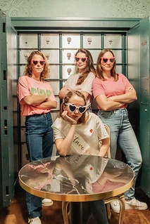 Quatre amies en t-shirts colorés et lunettes en cœur posent dans une salle de coffre pendant un escape game EVJF à Paris