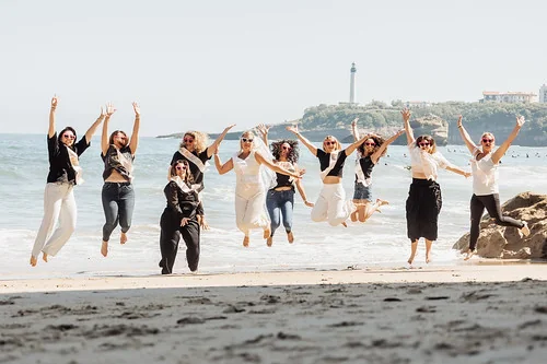 Groupe de femmes sautant sur la plage pendant un EVJF à Biarritz, avec vue sur l’océan et le phare en arrière-plan.