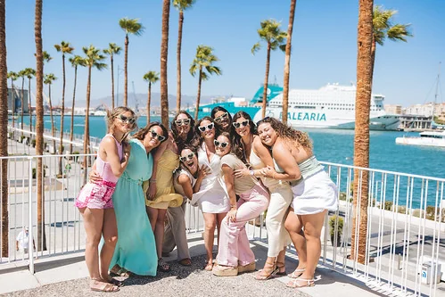 Groupe de femmes souriantes posant au port de Malaga pendant un EVJF ensoleillé, entourées de palmiers et vue sur la mer.