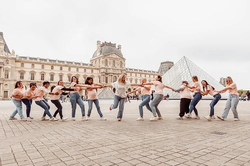 groupe de femmes en jeans et t-shirts roses jouant en chaîne humaine devant la pyramide du louvre pendant un evjf à paris