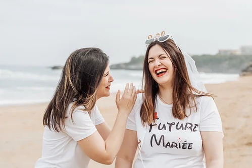 future mariee riant sur la plage de biarritz pendant un evjf tandis qu une amie lui chuchote a l oreille lors des gages et defis