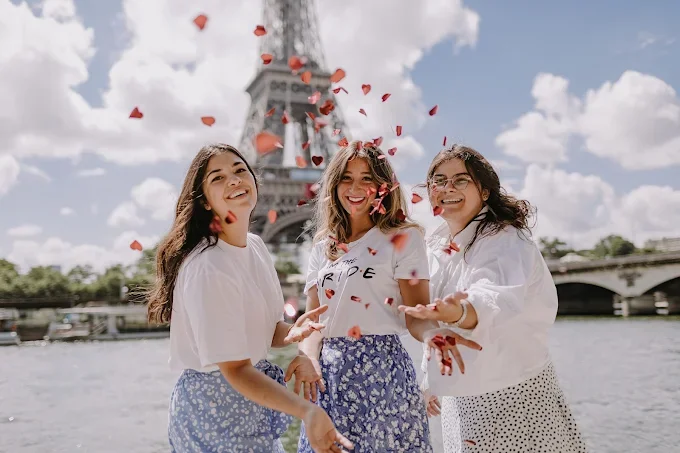 Trois femmes souriantes pendant un  lançant des pétales de fleurs devant la Tour Eiffel sous un ciel ensoleillé.