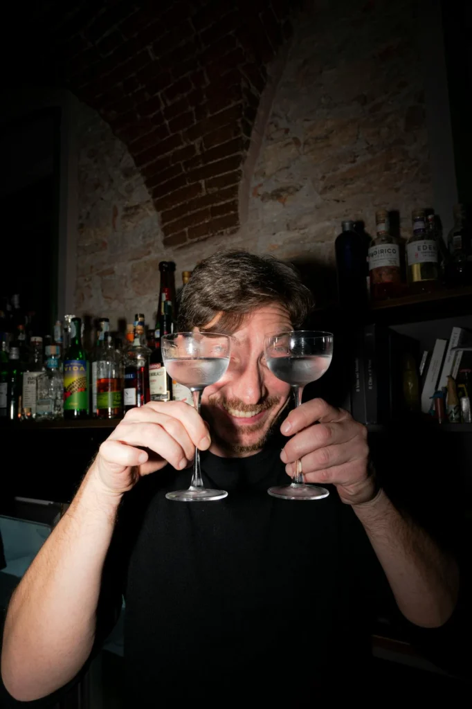 Homme souriant tenant deux verres à cocktail devant ses yeux dans un bar tamisé.